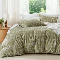 a bed with green comforter and pillows in front of a white curtained window