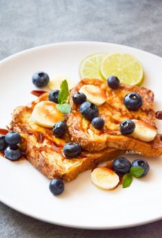 two pieces of french toast with blueberries and bananas on a white plate next to lemon wedges