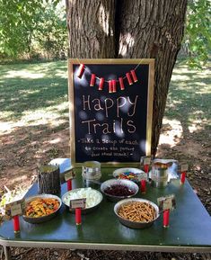 a picnic table set up with food and drinks for a happy trails party in the woods