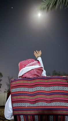 a man sitting on top of a colorful blanket under a moon lit sky with his hands in the air