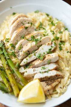 a white bowl filled with pasta and chicken next to asparagus, lemon wedges and parsley