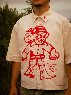 a man wearing a red and white shirt with a cartoon character on the chest, standing in front of trees