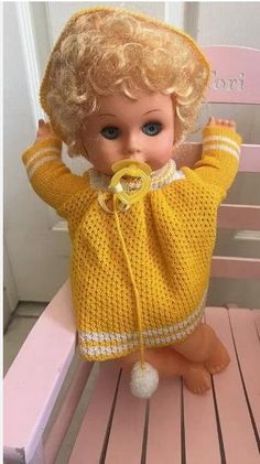 a doll sitting on top of a wooden bench wearing a yellow sweater and white shoes