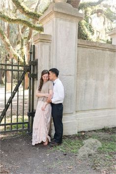 a man and woman standing next to each other in front of a fence with trees