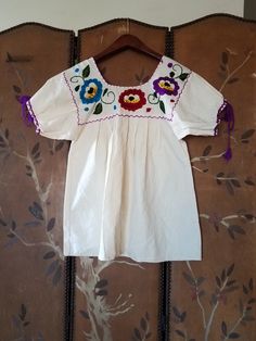 80s lovely ivory cotton Mexican hippie boho peasant blouse. Purple red and blue embroidered flowers with swirly green leaves. The square neck is edged in bright purple so are the cuffs of sleeves with long purple tassels Pleated front and back. Bust small 36 Across shoulders 13 Length 26 Summer Festival Peasant Top With Embroidered Sleeves, Bohemian Summer Peasant Top With Embroidered Sleeves, Bohemian Peasant Top With Embroidered Sleeves For Summer, Spring Festival Beige Peasant Top, Summer Cotton Peasant Top With Embroidered Sleeves, White Hippie Peasant Top For Festival, Fall Cotton Short Sleeve Peasant Top, Fall Cotton Peasant Top With Short Sleeves, Fall Short Sleeve Cotton Peasant Top