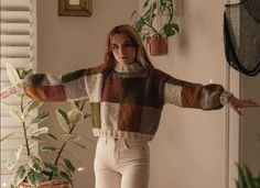 a woman standing next to a potted plant wearing a sweater and pants with her arms outstretched