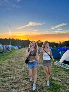 two young women walking in front of tents at an outdoor camping site with the sun setting behind them