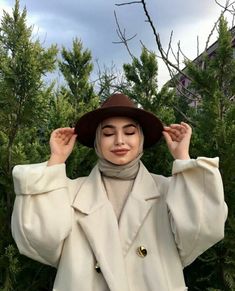 a woman wearing a white coat and brown hat standing in front of trees with her hands on her head