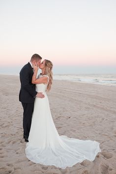 a bride and groom kiss on the beach at sunset in front of the ocean as the sun sets