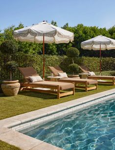 lounge chairs and umbrellas near the edge of a swimming pool with grass in the foreground