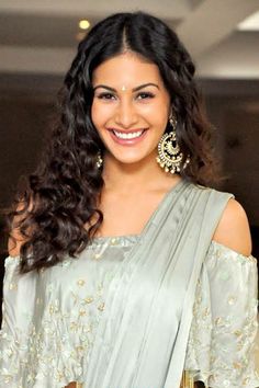 a woman with long dark hair wearing a grey sari and gold earrings smiles at the camera