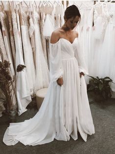 a woman in a white dress standing next to some dresses hanging on a rack and looking down at the floor