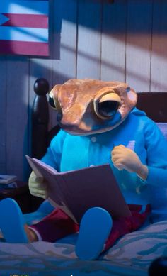 a stuffed frog reading a book on top of a bed in a room with blue walls