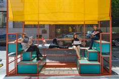 three people sitting on benches in front of a yellow and orange structure with an awning