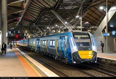 a blue and yellow train pulling into a station with people standing on the platform next to it