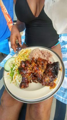 a woman holding a plate with meat and vegetables on it while sitting in a beach chair