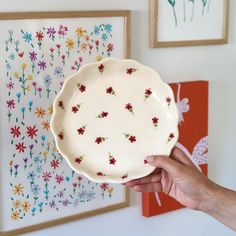 a hand holding a white plate with red and blue flowers on it in front of paintings