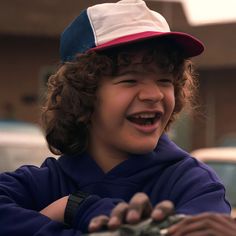 a young boy with curly hair wearing a blue and white baseball cap smiles at the camera