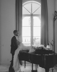 a man standing next to a grand piano in front of a large window with long white feathers on it