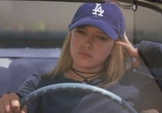 a woman sitting in the drivers seat of a car wearing a dodgers hat and holding her hand to her head