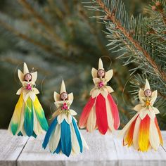 three different colored angel ornaments sitting on top of a wooden table next to a pine tree