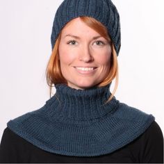 a woman with red hair wearing a blue knitted hat and turtle neck sweater smiles at the camera