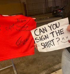 a woman holding up a sign that says can you sign the shirt?