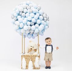 a little boy standing next to a teddy bear in a basket with blue and silver balloons