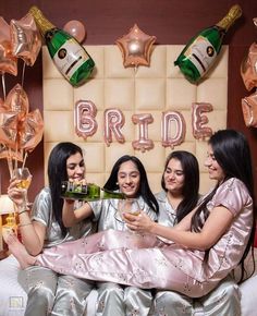 three women toasting champagne while sitting on a bed with balloons and streamers in the background