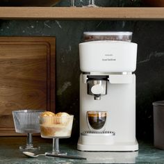 a white coffee maker sitting on top of a counter next to a cup filled with muffins