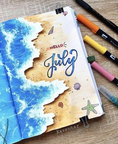 an open notebook with the words hello july written on it next to markers and pencils