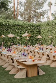 an outdoor dining table set up with umbrellas and flowers on the tables in front of a hedge