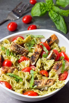 a white bowl filled with pasta salad and topped with grilled chicken, cherry tomatoes and basil