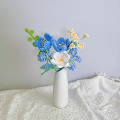 a white vase filled with blue flowers on top of a lace covered table cloth next to a gray wall