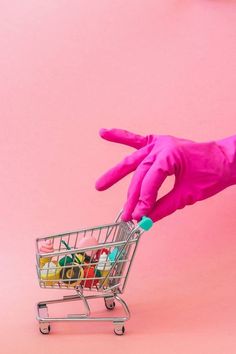 a hand in pink gloves pushing a shopping cart filled with toys on a pink background