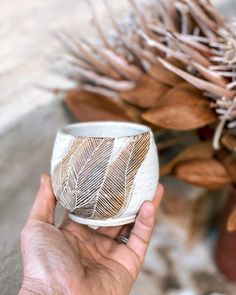 a hand holding a white and gold cup with leaves on the inside, in front of some dried plants