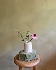 a white vase with pink flowers sitting on top of a wooden stool next to a wall