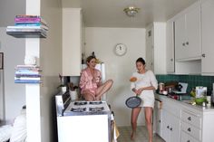 two women sitting on the counter in a small kitchen, one holding a frying pan