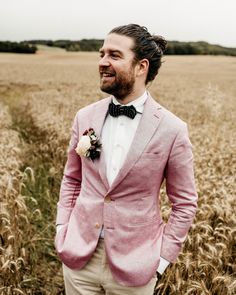 a man in a pink suit and bow tie standing in front of a wheat field