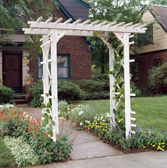 a white wooden arbor in front of a house with flowers and plants growing around it