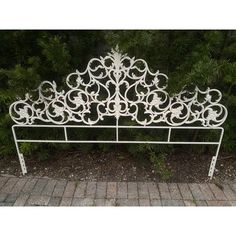 a white iron bed frame sitting on top of a brick walkway next to trees and bushes