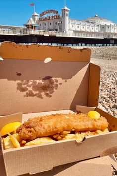 fish and chips in a cardboard box on the beach with buildings in the back ground