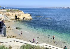 people are standing on the beach near the water and rocks, while others walk along the shore