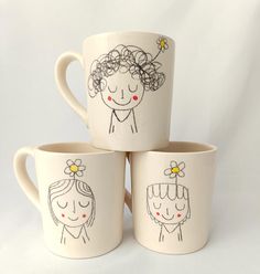 three coffee mugs with hand drawn faces and flowers on the sides, one has a flower in her hair