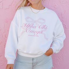 a woman standing in front of a pink wall wearing a white sweatshirt with the words, apha oh omega on it