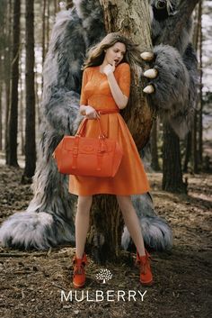 a woman in an orange dress is standing next to a large gray bear and holding a red handbag