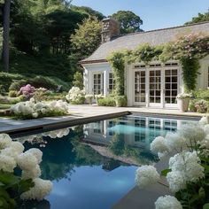 a pool surrounded by white flowers next to a house with large windows and lots of greenery