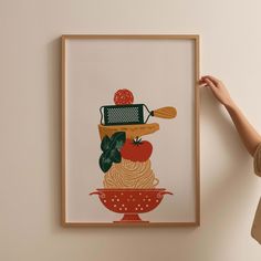 a woman is holding up a framed art print with vegetables and pasta on it in front of a wall