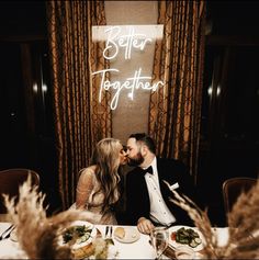 a man and woman sitting at a table with food in front of them, kissing