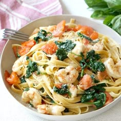 a white bowl filled with pasta and shrimp on top of a table next to basil leaves